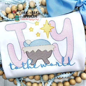 May include: Embroidered design of the word "Joy" in pink with a manger scene and a star in the center. The words "to the world" are embroidered in blue below the word "Joy".