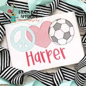May include: White t-shirt with embroidered peace sign, heart, and soccer ball. The name "Harper" is embroidered in pink. The shirt is surrounded by black and white striped ribbon and light blue ribbon.