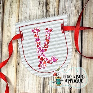 Puede incluir: Un banderín de tela con un borde cosido rojo y una letra "V" llena de patrones de corazones. El banderín es de rayas grises y blancas, con cinta roja adjunta para colgar. El banderín tiene el texto "HUG A BUG APPLIQUE".