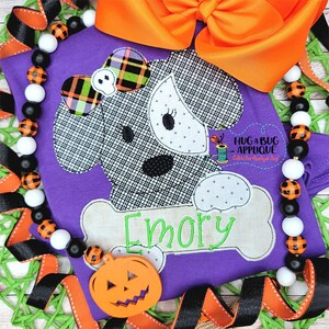 May include: Purple shirt with a black and white dog applique wearing a plaid bow. The dog is holding a bone with the name "Emory" embroidered on it. A pumpkin and a black and white beaded necklace are on the shirt.