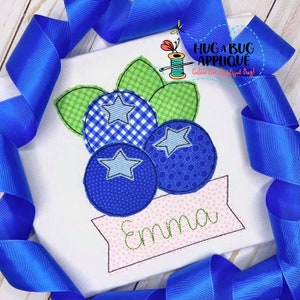 May include: Embroidered design of three blue blueberries with green leaves and a pink banner with the name "Emma" stitched on it.