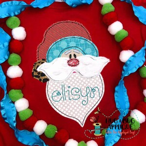 May include: A red, white, and blue applique of a Santa Claus face with the name "ellison" embroidered on the beard. The applique is on a red shirt with a blue ribbon and a garland of red, white, and green pom-poms.