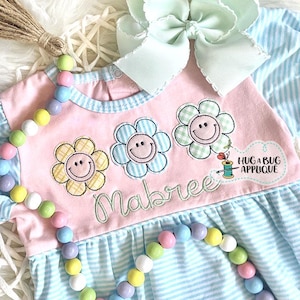 May include: A pink and blue striped baby dress with a pink bodice and three embroidered yellow, blue, and green flowers with smiley faces. The dress has the name "Mabriee" embroidered on the bodice. The dress is decorated with a string of pastel colored beads.