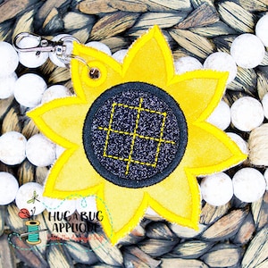 Könnte beinhalten: Gelbe Sonnenblumen-Applikation mit schwarzem Mittelteil und gelben Nahtdetails. Die Applikation hat einen silbernen Verschluss. Der Text "HUG A BUG APPLIQUE" ist in der linken unteren Ecke sichtbar.