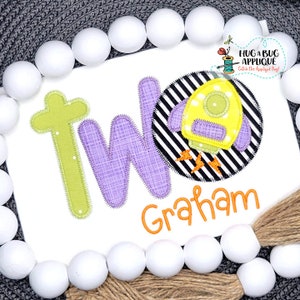 May include: A white onesie with a purple and green applique of the word "TWO" and a yellow rocket ship with black and white stripes. The name "Graham" is embroidered in orange below the applique.