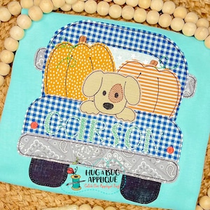 May include: A blue and white gingham truck with a gray paisley trim. The truck is carrying two orange pumpkins and a brown puppy with a white chest. The truck has the name "GIBSON" embroidered on the back.