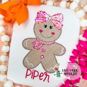 May include: A white t-shirt with a gingerbread person applique. The gingerbread person has a pink bow, scarf, and rosy cheeks. The name "Piper" is embroidered in pink script. A logo for "HugaBug Applique" is in the lower right corner.