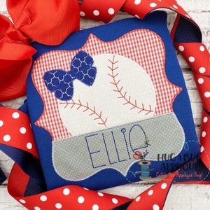 May include: A blue and white baseball-themed appliqué with a red and white gingham border. The appliqué features a baseball with stitching, a blue bow, and the name "Ellie".