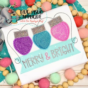 May include: White shirt with an embroidered design of three light bulbs with a banner that reads "Merry & Bright".