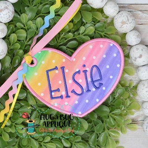 May include: A heart-shaped, rainbow-colored fabric patch with white polka dots and the name "Elsie" embroidered in purple thread.