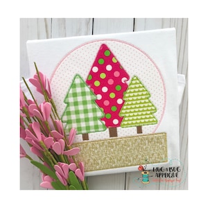 May include: A white t-shirt with a green, pink, and white applique of three Christmas trees in a circle. The trees are surrounded by a pink and white polka dot background. The bottom of the design is a brown and gold fabric.