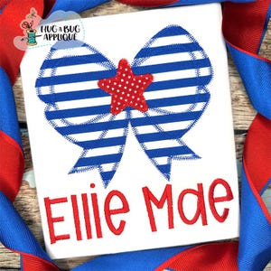 May include: White t-shirt with a patriotic design featuring a blue and white striped bow with a red star in the center. The name "Ellie Mae" is embroidered in red script below the bow. The design is framed by red and blue ribbon.