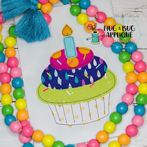 May include: A colorful embroidered cupcake applique with a blue candle, pink and yellow frosting, and sprinkles. The applique is on a white fabric background.