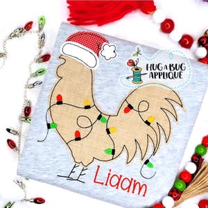 May include: A gray shirt with a brown rooster wearing a red and white Santa hat with a white brim. The rooster is decorated with a string of colorful Christmas lights. The shirt is personalized with the name "Liam".