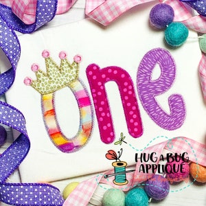 May include: A pink, purple, and gold applique design of the word "one" with a crown on top. The design is on a white fabric background. The text "Hug a Bug Applique" is visible in the bottom right corner.