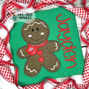 May include: A green shirt featuring a gingerbread man design. The gingerbread man is brown with a red scarf and buttons. The name "Jayden" is embroidered in red. The shirt is framed by red and white checked ribbon.