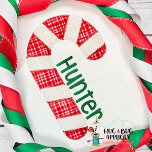 Puede incluir: Un pelele blanco con una aplicación de bastón de caramelo rojo y blanco y el nombre "Hunter" bordado en verde. El pelele está rodeado de cinta roja, blanca y verde.