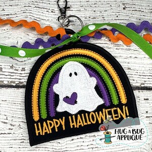 May include: A black Halloween-themed keychain with a white ghost design, a purple heart, and a rainbow. The text "HAPPY HALLOWEEN!" is in gold. The keychain has a silver clasp and is decorated with orange, purple, and green ribbons.