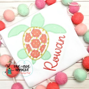 May include: A white t-shirt with a coral and green applique of a sea turtle. The name "Rowan" is embroidered in coral thread below the turtle.