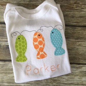 May include: White baby onesie with three embroidered fish in green, orange, and blue with white polka dots. The fish are connected by a black string. The name "Parker" is embroidered below the fish.