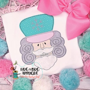 以下が含まれることがあります： くるみ割り人形の顔のアップリケが付いた白いTシャツ。デザインには、雪の結晶が付いたターコイズの帽子、白い口ひげとあごひげ、そして「HUG A BUG APPLIQUE Catch The Applique Bug！」というテキストが含まれています。