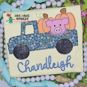 Może przedstawiać: Niebieski samochód dostawczy z kwiatowym wzorem, przewożący dwie pomarańczowe dynie i różową świnię. Samochód ma czarne koła, a pod nim wyszyte jest imię "Chandleigh" niebieską nicią.