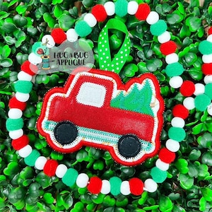 Puede incluir: Camión rojo de fieltro con un árbol de Navidad verde de fieltro en la parte trasera. El camión está decorado con costuras blancas y ruedas de fieltro negro. El camión está colgado de una cinta de lunares verde y blanca.