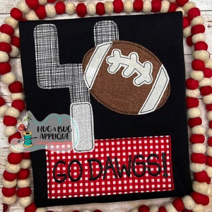 May include: Black t-shirt with a white and gray football and goal post design. The shirt also has a red and white checkered rectangle with the text "GODAWGS!" in black.