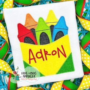 May include: A yellow box with a green and yellow lid filled with five colorful crayons. The box has the name "Adron" written in red on the front.