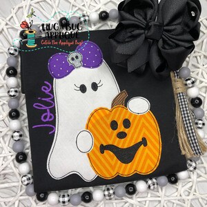 May include: A black t-shirt with a white ghost holding a pumpkin. The ghost has a purple bow with a skull and crossbones on it. The pumpkin has a smiling face. The text "Jolie" is embroidered on the shirt.
