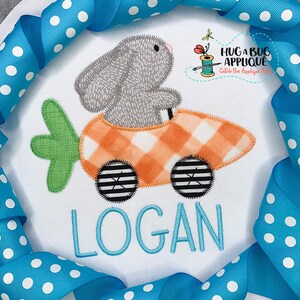 May include: A grey bunny driving a carrot-shaped car with black and white wheels. The car is orange and white checked. The bunny is driving on a white background with the name "LOGAN" embroidered in blue below the car.
