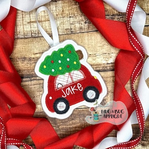 Puede incluir: Coche rojo de fieltro con un árbol de Navidad verde de fieltro en la parte superior. El coche tiene una ventana de fieltro blanco y el nombre "Jake" está bordado en el lateral.