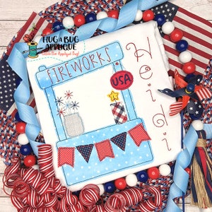 May include: A white fabric square with a blue and red fireworks stand design. The stand has a banner that says "FIREWORKS" and a "USA" sign. The design includes fireworks, stars, and red, white, and blue bunting.
