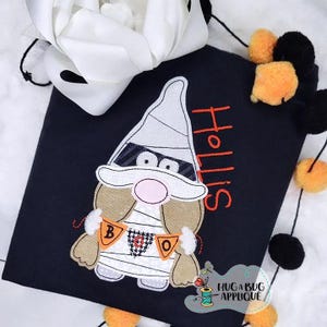 Op de afbeelding: Een zwart T-shirt met een mummie-dwerg-applicatie. De dwerg draagt een wit mummie-kostuum met een zwart oogmasker en houdt een "BOO"-banner vast. De naam "Hollis" is in rood op het shirt geborduurd.