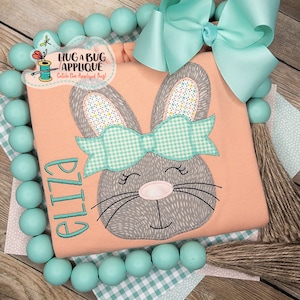 May include: A peach-colored shirt with a gray bunny applique wearing a green and white gingham bow. The bunny has a pink nose and closed eyes. The shirt has the name "Eliza" embroidered on the front.