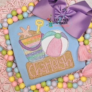 May include: A light blue t-shirt with an embroidered design of a beach bucket, shovel, and beach ball. The name "Charleigh" is embroidered below the design.