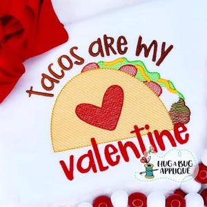 以下が含まれることがあります： 白い生地に刺繍されたテキストとタコスデザイン。「Tacos are my Valentine」というテキストが茶色と赤で書かれています。タコスには赤いハートとカラフルな具材があります。「HUG A BUG APPLIQUE」のロゴが右下にあります。