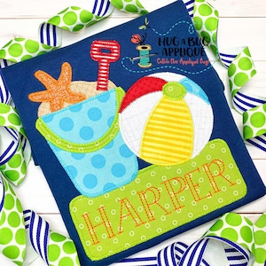 May include: A blue t-shirt with a green, yellow, red, white, and orange applique of a beach bucket, shovel, starfish, and beach ball. The name "HARPER" is stitched in white on a green rectangle.