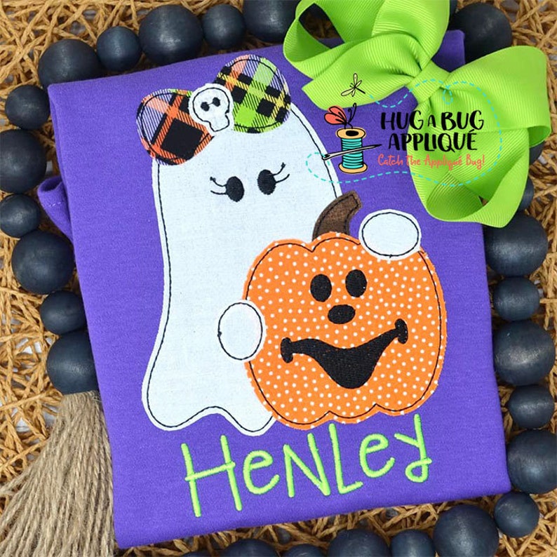 May include: Purple shirt with a white ghost holding an orange pumpkin with a black face. The ghost has a plaid bow and the shirt says "Henley".
