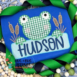 May include: A blue applique design featuring a green frog with a gingham pattern, surrounded by brown reeds. The name "HUDSON" is stitched in blue on a white and blue striped banner.