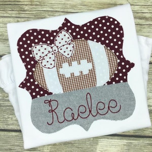 May include: A white t-shirt with a maroon and white football and bow applique. The applique is surrounded by a maroon and white polka dot frame. The name "Raelee" is embroidered in gray on a gray banner.