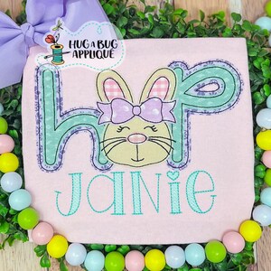 May include: A pink shirt with the word "Hop" embroidered in green and blue with a bunny head in the middle. The bunny has a pink bow and pink and white checkered ears. The name "Janie" is embroidered below in green.