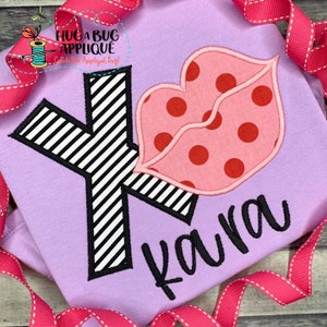 Puede incluir: Una camiseta morada con una letra K a rayas blancas y negras y una aplicación de labios rosa y rojos. El nombre "Kara" está bordado en negro debajo de la letra.