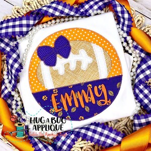 May include: A white t-shirt with a purple, orange, and brown applique of a football with a bow on top. The applique is surrounded by a circle of orange and white polka dots. The name "Emmy" is written in orange on a purple background with a paw print pattern.