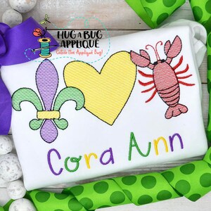 May include: A white shirt with embroidered images of a fleur-de-lis, a heart, and a crawfish. The name "Cora Ann" is embroidered below the images. The text "Hug a Bug Applique" is also embroidered on the shirt.