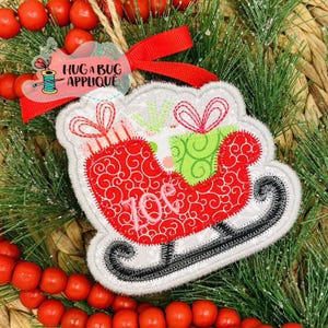 May include: A white felt Christmas ornament with a red and green embroidered sleigh. The sleigh is filled with presents and the word "Joy" is embroidered on the side.