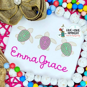 May include: White t-shirt with the name "Emma Grace" embroidered in pink cursive lettering. Three embroidered sea turtles with green and pink shells are above the name. The text "Hug a Bug Applique Catch the Applique Bug!" is in the top right corner.