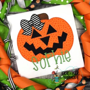 Pode incluir: Uma camiseta branca com uma aplicação de abóbora laranja e preta com um laço listrado preto e branco. O nome "Sophie" está bordado em verde abaixo da abóbora. A camiseta é decorada com fita laranja e verde.