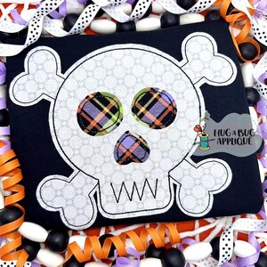 May include: White skull and crossbones applique with plaid fabric accents in shades of pink, orange, green, and purple. The applique is on a black fabric background. The text "HUG A BUG APPLIQUE" is visible in the bottom right corner.