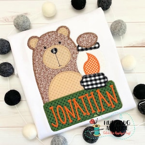 May include: A brown bear applique with a campfire and a green banner with the name "Jonathan" in orange text.